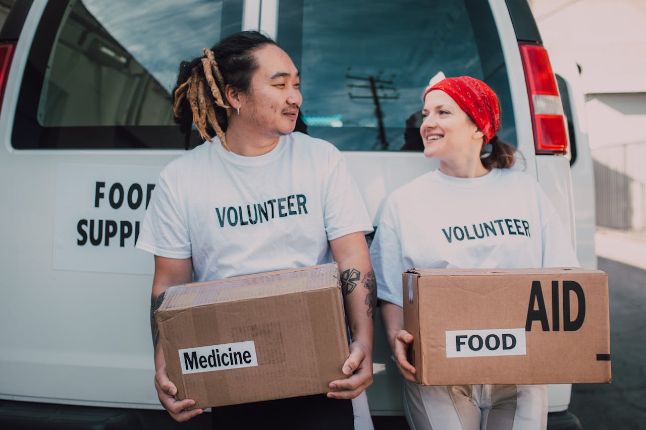 Two smiling volunteers carrying boxes labeled food and medicine by a supply van.